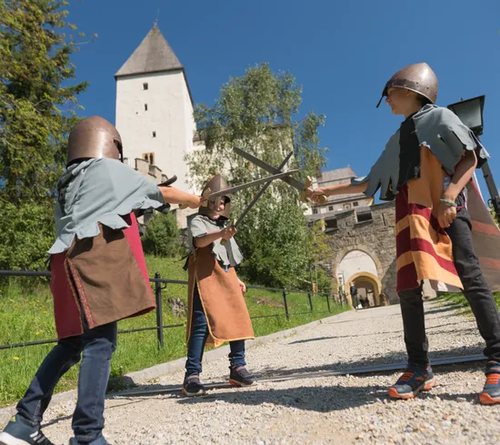 Kinder in mittelalterlichen Kostümen spielen Schwertkampf vor einer historischen Burg bei Sonnenschein